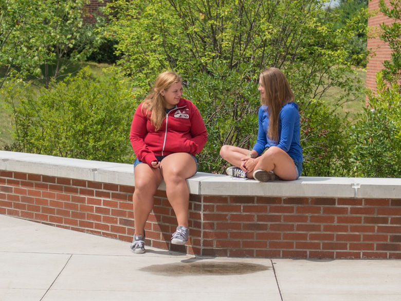 two high school students sit on a brick bench surrounded by trees