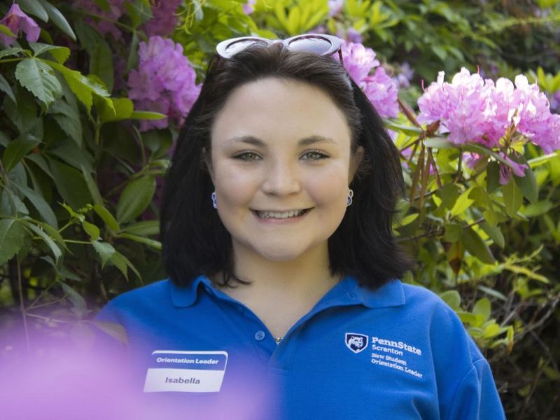 Student smiles in front of flowers wearing a shirt that reads 'New Student Orientation'