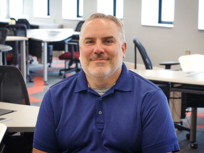 photo of Rob Notari sitting in IT lab with computers and windows behind him