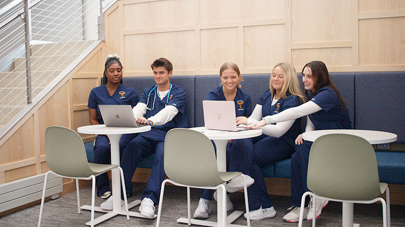 5 students in scrubs use laptops at a comfortable looking seating at tables in a brightly lit area