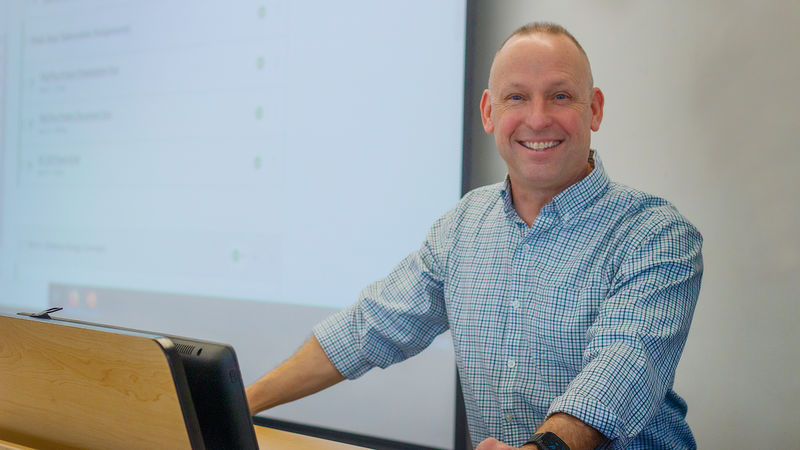smiling man behind a podium and computer monitor in from of screen displaying tech class information
