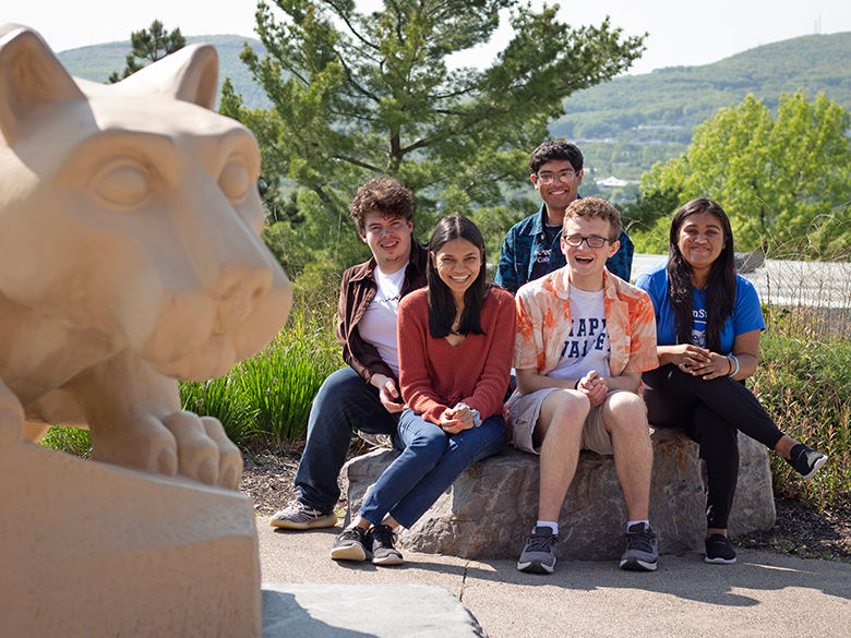 four smiling studnets sit near the penn state scranton lion shrine statue that overlooks a mountain and valley