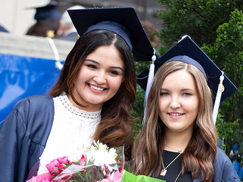 two smiling females in graduation cap and gowns holding flowers