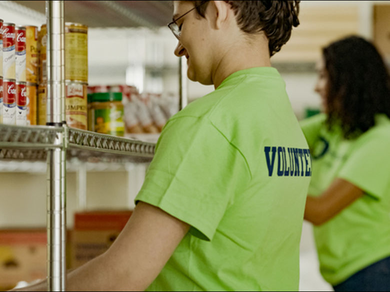 two students wearing volunteer tee shirts stock canned goods at food pantry