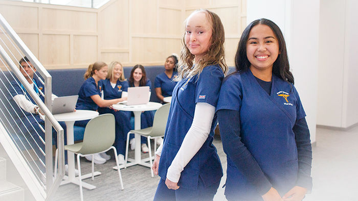 two smiling nursing students back to back with a group of nursing students on computers in the background