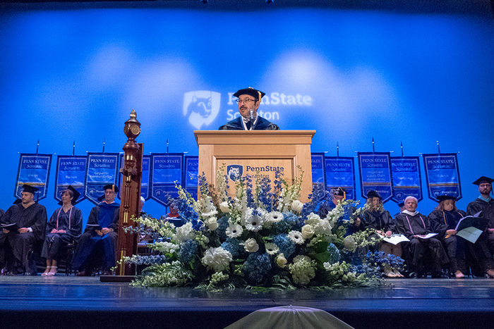 Man wearing graduation hat and robe on stage in front of a podium. Stage party, people  seated behind him.