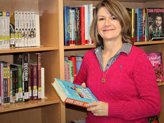 Mary Ann Joyce holding a book in front of the non-fiction collection in the library