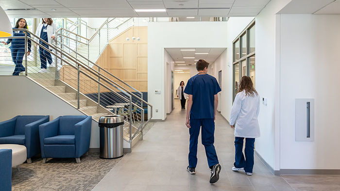 students wearing scrubs walk along a long brightly lit hallway and descend a large modern stair case