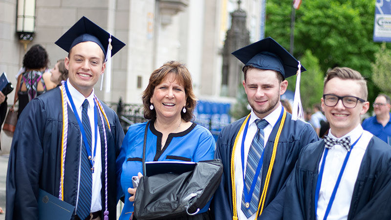 three graduates in cap and gowns pose with faculty member
