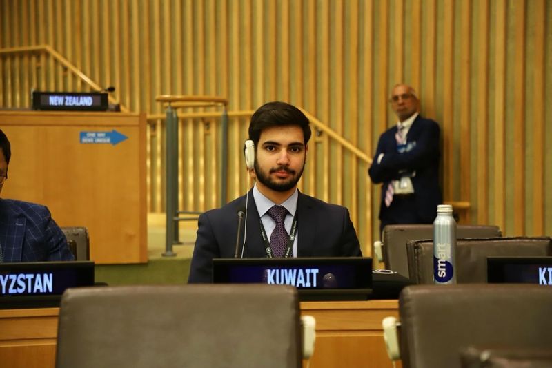 A student wearing a suit and headphones sits with a sign reading "Kuwait" in front of him