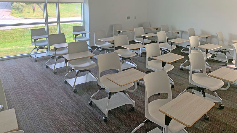white chairs with attached desktop in a large classroom with large window and shades