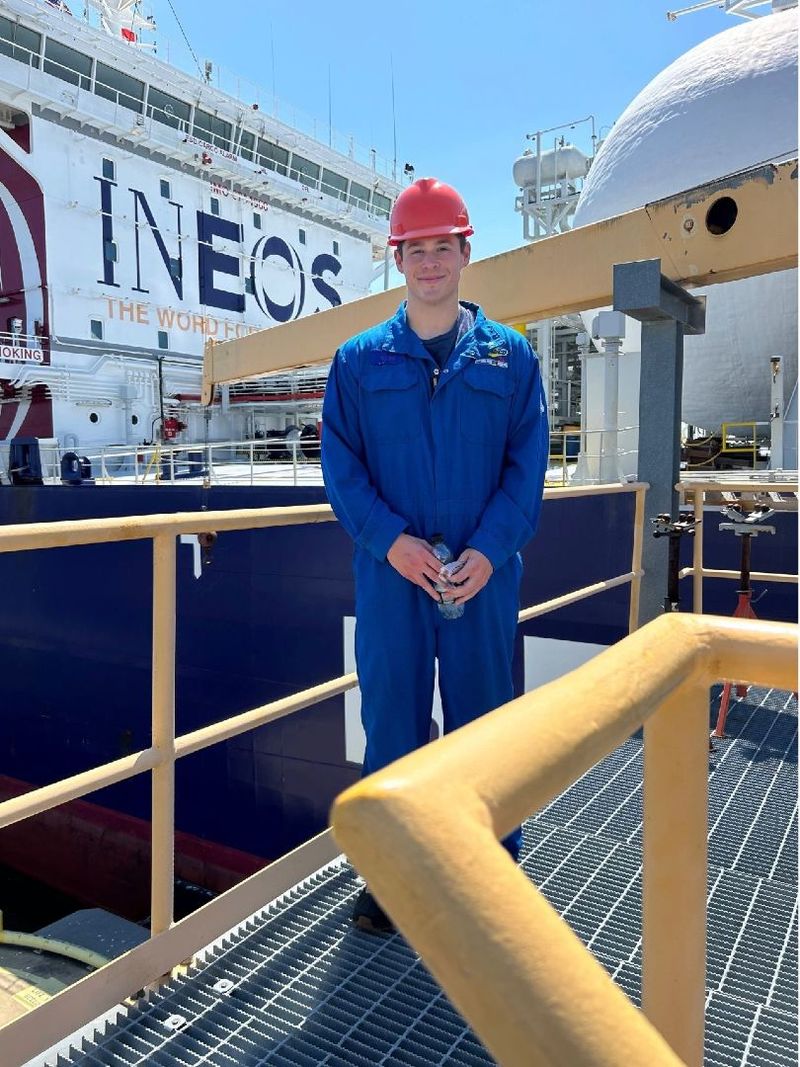 A student wears blue coveralls and an orange hard hat on a job site