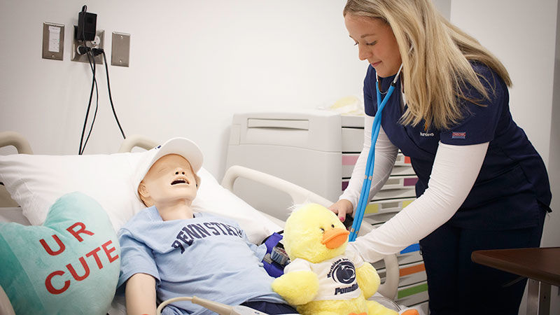 student in nursing scrubs with stetescope simulates taking blood pressure of a child mannequin in a lab that looks like a hospital room