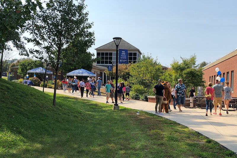 large group of people walk on sidewalk through across green grass campus with buildings in the background.