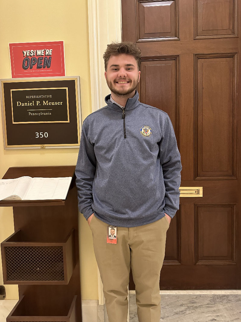 A student stands in front of an office with a sign that reads "Representative Daniel P. Meuser" 