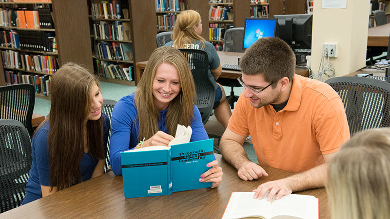 Three students share a textbook at a library table.