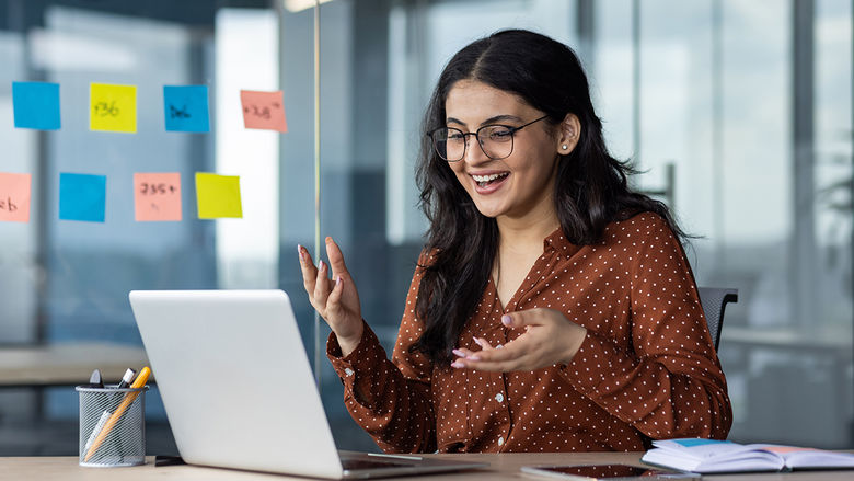 a student at a laptop smiling and gesturing to sticky notes on a whiteboard