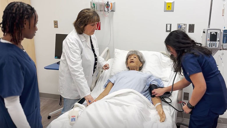 woman in white lab coat works on a patient simulator mannequin with two students in a hospital room