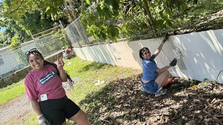 Three female students work on painting an outside garden wall