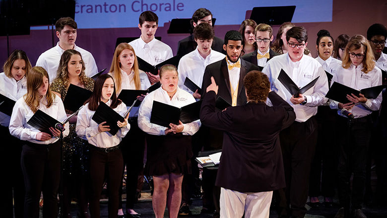 group of about twenty students holding music books being led in song by their music director