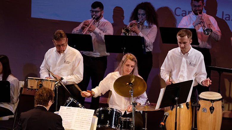 3 people in drum section in a jazz band. # hor players behind them