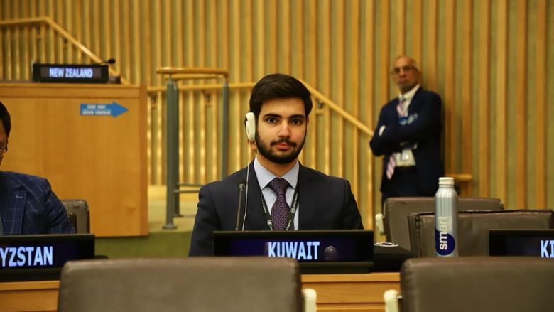 A student wearing a suit and headphones sits with a sign reading "Kuwait" in front of him