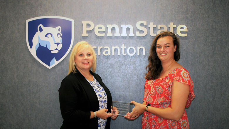 Chris Ostroski handing Katelin McAndrew her glass Ridge View Society plaque in front of the Penn State Scranton logo wall