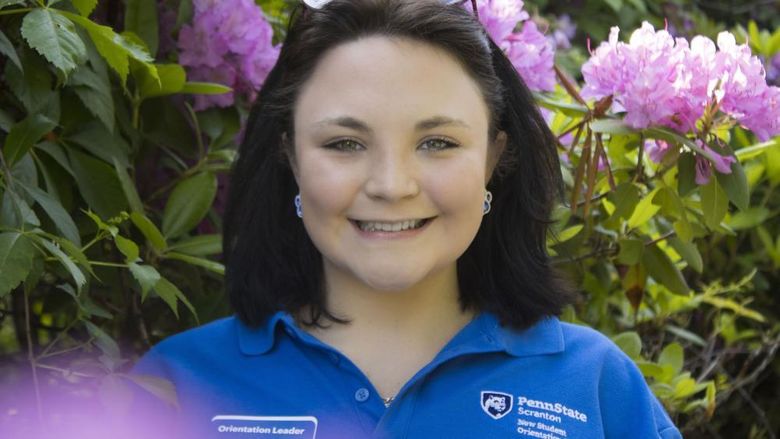 Student smiles in front of flowers wearing a shirt that reads 'New Student Orientation'
