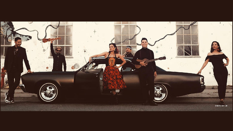 Members of a band hold instruments while standing around a black car