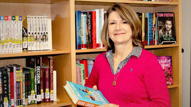 Mary Ann Joyce poses for a photo with a book, with a multi-shelf bookcase full of books in background.
