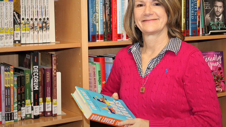 Mary Ann Joyce holding a book in front of the non-fiction collection in the library
