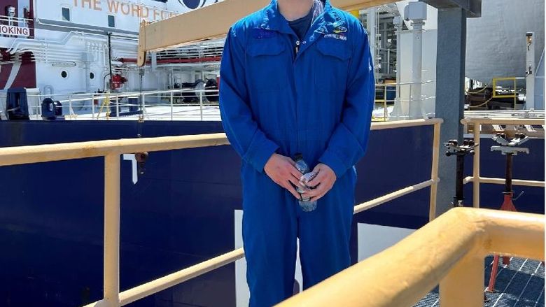 A student wears blue coveralls and an orange hard hat on a job site