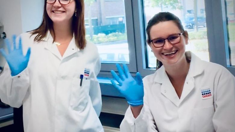 Two students wearing lab coats and blue rubber gloves smile for the camera