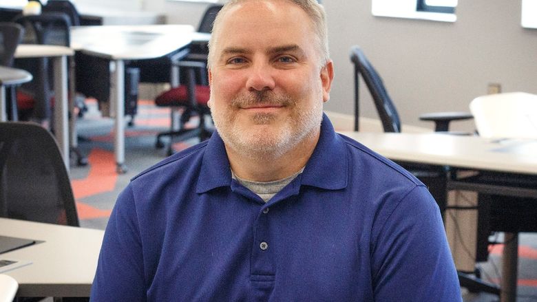 photo of Rob Notari sitting in IT lab with computers and windows behind him