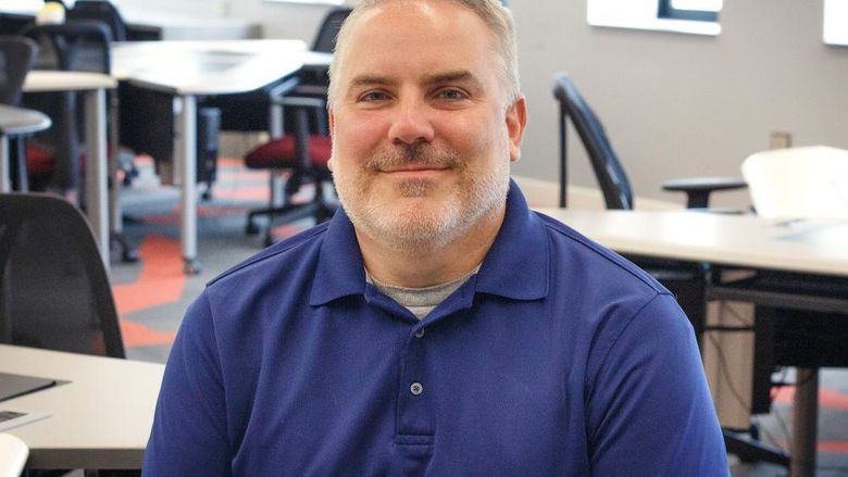 photo of Rob Notari sitting in IT lab with computers and windows behind him