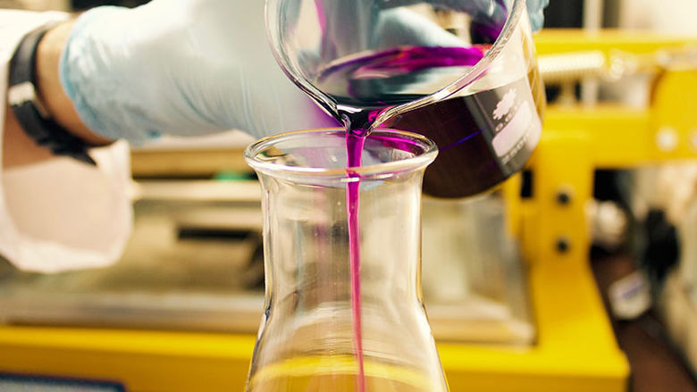 hand with latex glove pours purple liquid into a glass beaker