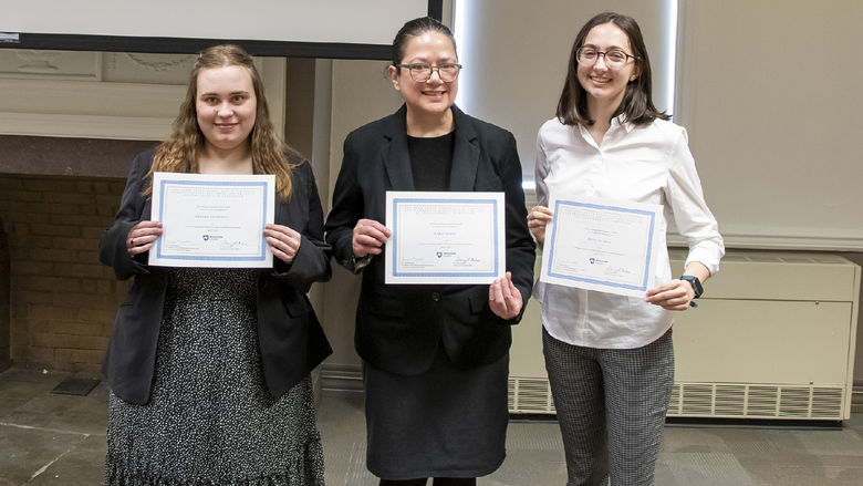 Three students stand in front of a fire place with certificates