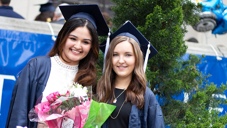 two girls at graduation wearing grad cap and gown holding flowers