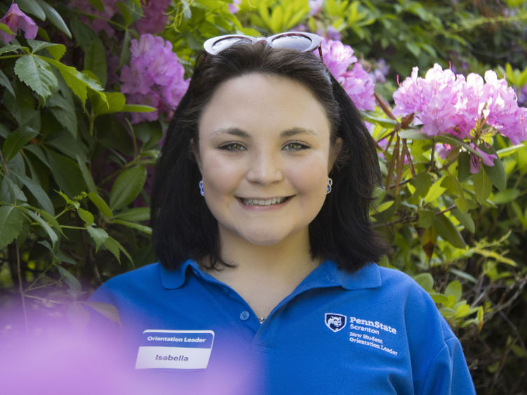 Student smiles in front of flowers wearing a shirt that reads 'New Student Orientation'